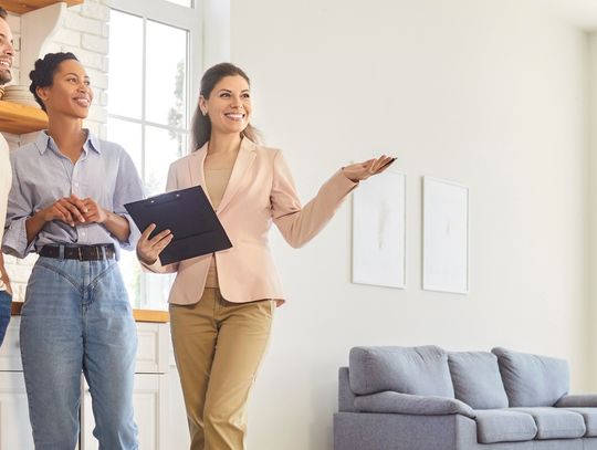 A female realtor holds a clipboard while showing the interior of a home to a young man and woman.