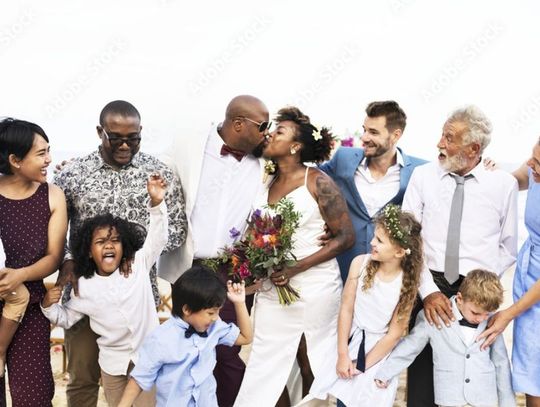 An African-American couple kiss on their wedding day. Their family surrounds them, smiling and cheering.