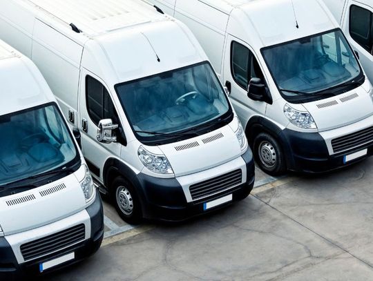A fleet of four identical service vans sits in a paved parking lot. The white vans are parked in an orderly row.