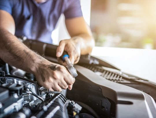 A close-up of a man in a T-shirt leaning over a car's engine and using a wrench on an exposed part.