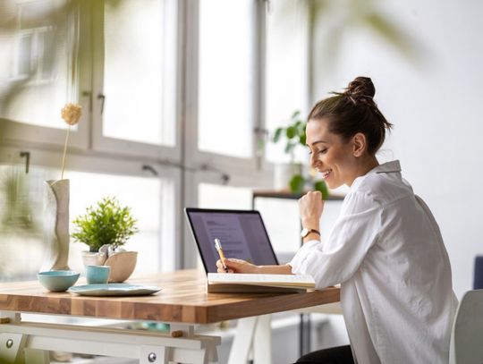 A woman in a white button-down shirt smiles as she writes in a notebook beside an open laptop on a wood desk.