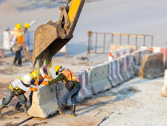 A group of construction workers wearing safety vests and hard hats using an excavator to lift a concrete barrier.