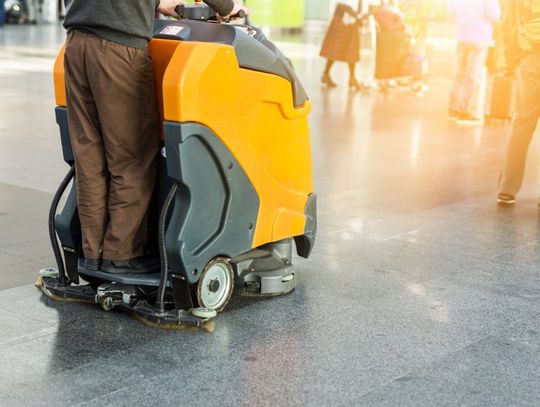 A custodian operating an industrial ride-on yellow floor cleaner across an expanse of tiled floor.