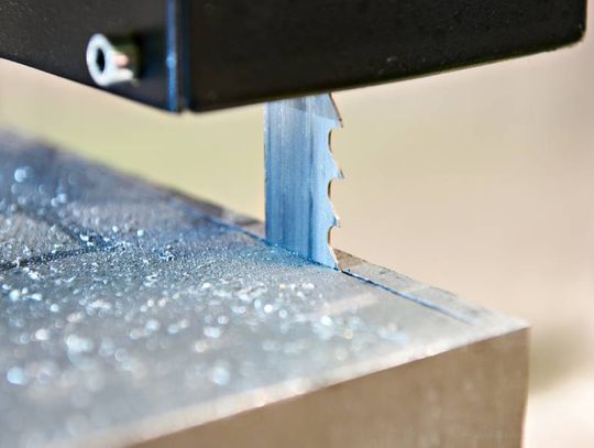 A close-up of a vertical band saw cutting a piece of metal. There are small metal shavings surrounding the blade.