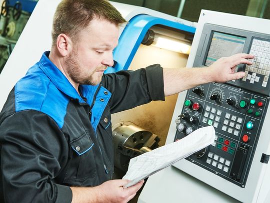 A machine operator is reading a manual while pressing machine buttons. He wears a black and blue shirt.