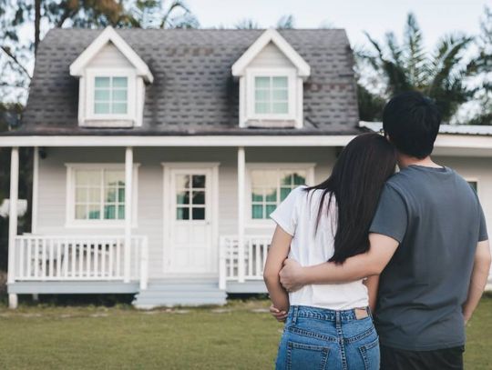 A young couple holding each other as they stand in their yard and face their white tiny home.