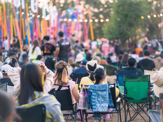 An overhead view of a crowd of people sitting on folding chairs in an outdoor area decorated with lights and streamers.
