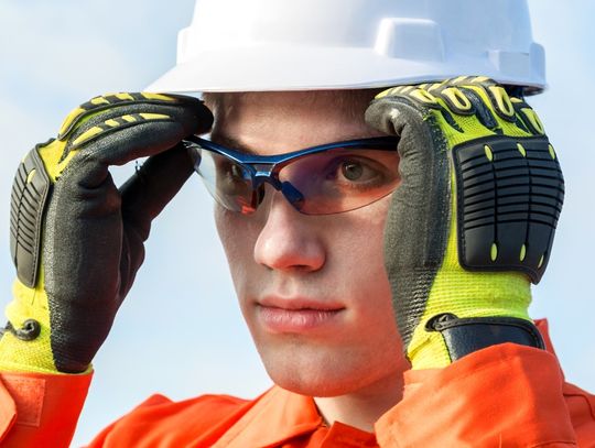 A young man in a white helmet and black and neon green work gloves is putting on blue safety goggles.