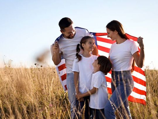 A family of four is standing outside on a sunny day. They are holding a large American flag over their shoulders.