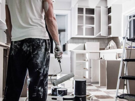 A male homeowner, covered in paint and holding a paint roller, standing in front of his kitchen in between renovations.