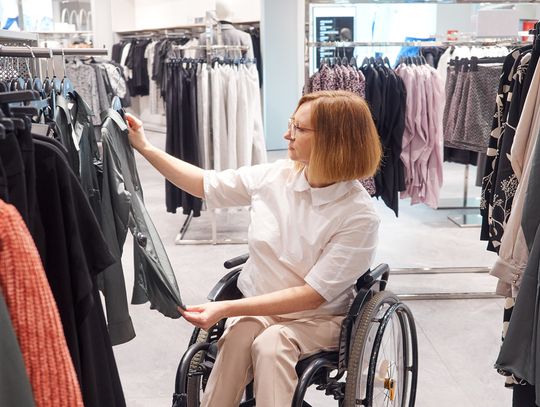 A female in a wheelchair is shopping in a clothing store. She is holding up a gray shirt on a hanger.