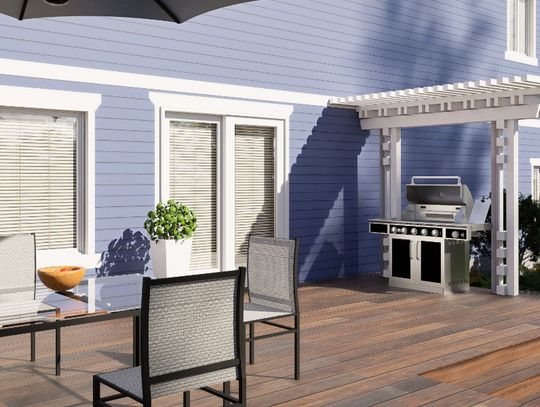 The deck in the backyard of a home with a blue house has a dining table, grill, pergola, and umbrella.