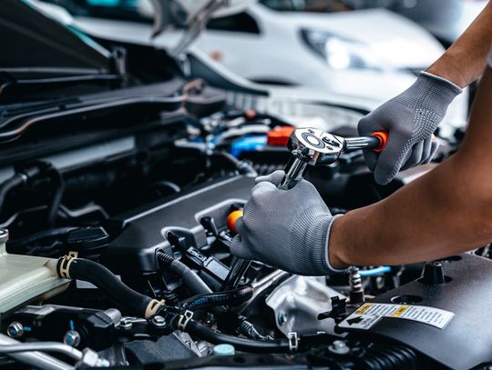 An individual in a gray T-shirt and gray gloves uses a wrench with an orange handle to work on a vehicle engine.