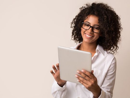 A woman with black curly hair standing against a beige background and smiling while looking at a silver tablet in her hands.