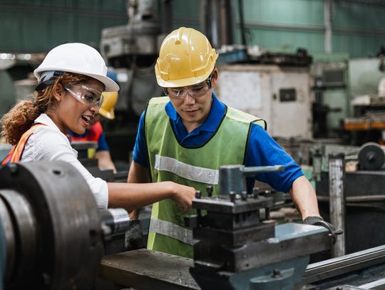 A man and a woman stand in front of a machine in a factory. Both wear hard hats, safety glasses, and safety vests.