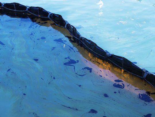 Floating containment boom on water with visible oil sheen, showing pollution control barrier across surface.