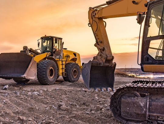 A yellow bulldozer and excavator working on a patch of furrowed, rocky dirt. The sky is orange.