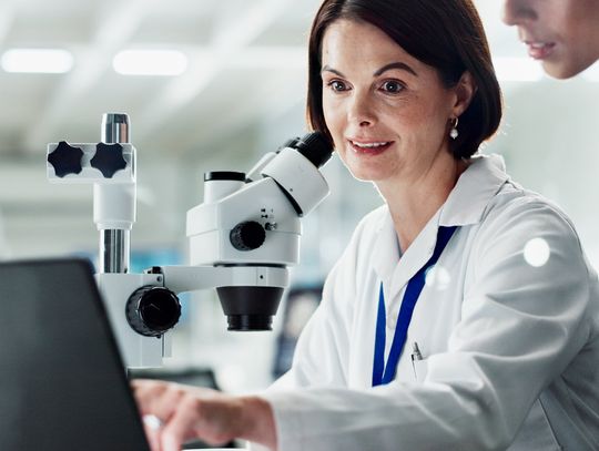 A scientist sits in a laboratory next to a microscope and points to a laptop screen while another scientist looks on.