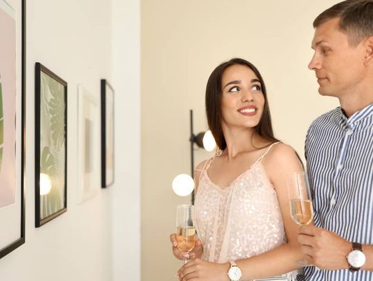 A young couple looking at each other and holding champagne flutes as they stand in front of a gallery exhibition.