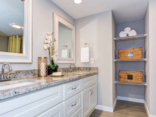 A home's bathroom with a granite countertop, two sinks, two mirrors, recessed shelving, and brown laminate flooring.