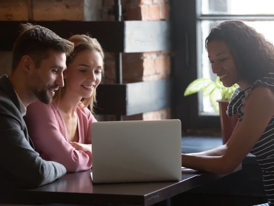 A woman sits with a couple at a table and shows them something on a laptop. What they are seeing makes them happy.