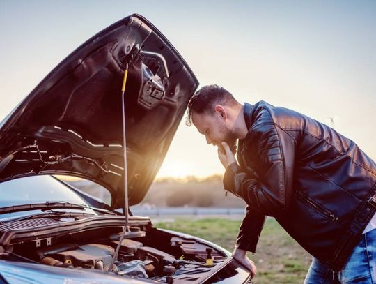 A young man in a leather jacket is looking at the engine of his car under the hood. There is a sunset in the background.