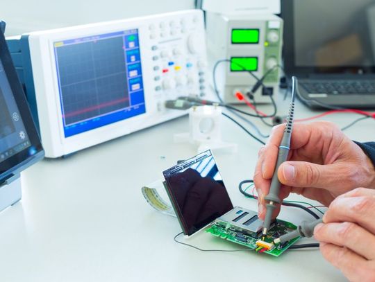 A technician's hands calibrate a piece of technology equipment on a table. A laptop lies on the surface nearby.