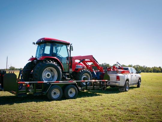 A white pickup truck tows a large red tractor with a black front bucket on a trailer parked on patchy grass.