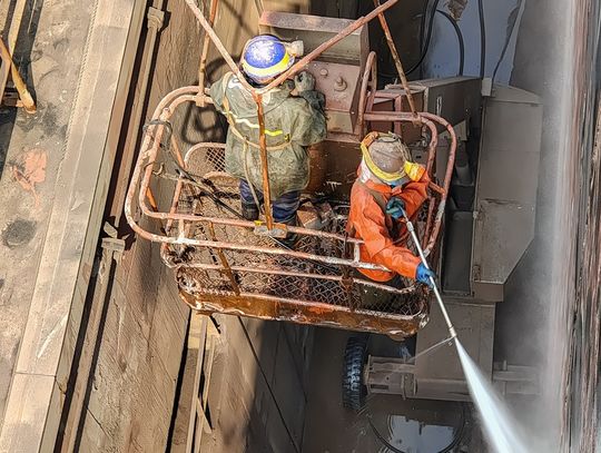 Two people wearing protective gear are in a boom lift and using a hydroblasting machine to clean a surface.