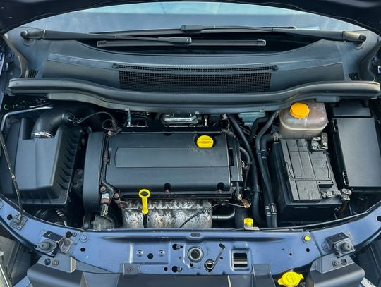 Overhead view of a car engine bay with a black plastic engine cover, yellow oil cap, battery, and coolant reservoir.