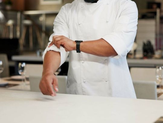 A man in a white chef’s jacket rolls up his sleeve while standing behind a white countertop in a dining area.