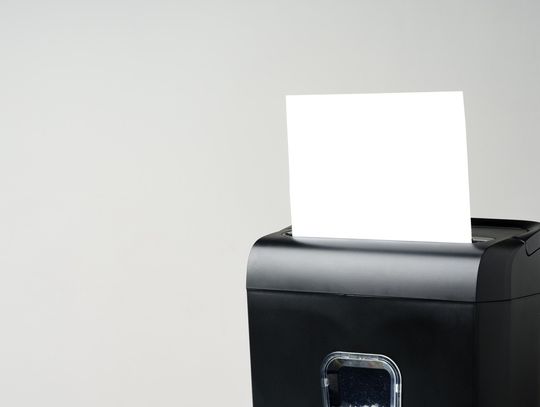 A blank piece of white paper sticks out of the top of a black shredder, which sits against a gray background.
