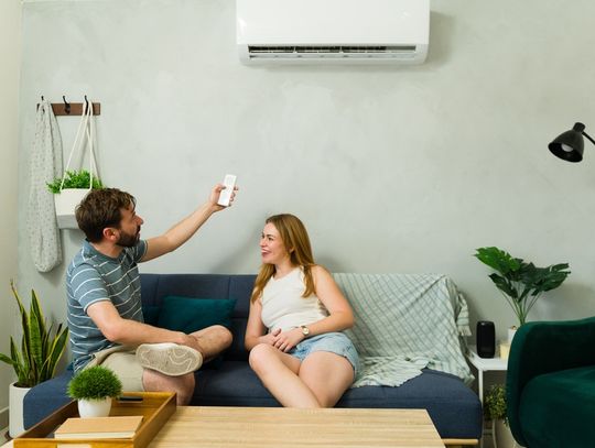 A man in a striped shirt holding an air conditioner remote while sitting beside a woman on a blue sofa in a living room.