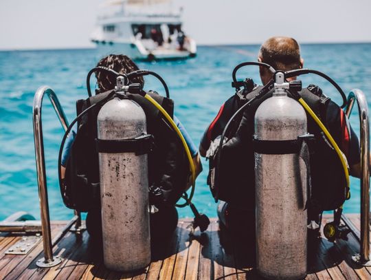 Two scuba divers sit on the edge of a wooden boat platform with their tanks and equipment, facing the blue ocean.