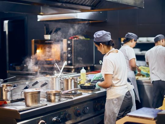 Several people in chef coats stand with their backs turned while working at counters in a commercial kitchen.