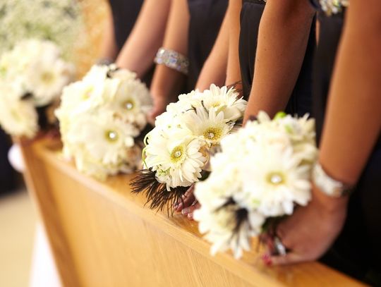 Bridesmaids in black dresses hold bouquets of white daisies with yellow centers while standing beside a wooden pew.