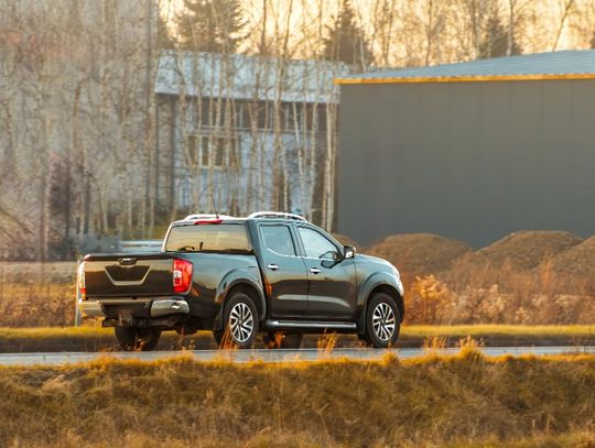 A wide view of a black pickup truck driving on a wet road past dry grass, leafless trees, and industrial buildings.
