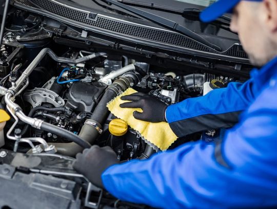 A mechanic in a blue uniform working under the hood of a car to diagnose and correct an issue with the powertrain system.