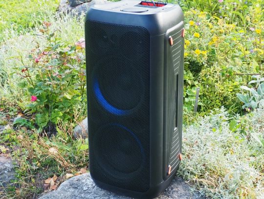 A black rectangular speaker sitting on a rock with grass and colorful wildflowers in the background.