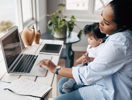 A mother holds her child in her lap as she talks on the phone and types on the computer.
