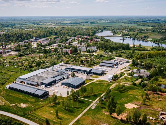 Aerial view of a small town surrounded by green fields, with industrial buildings and a lake nearby.