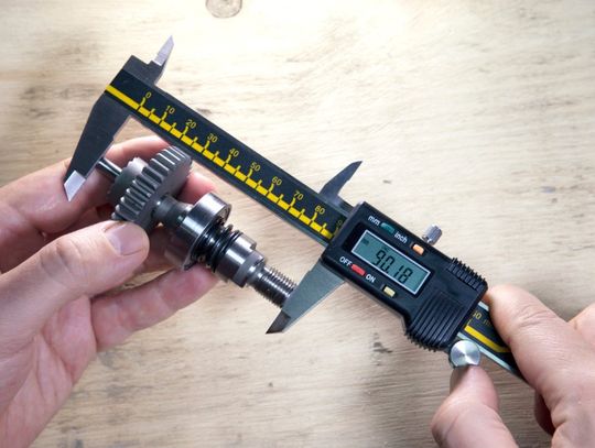 A machinist sitting at a wooden work table holding a digital caliper in both of their hands. The caliper reads 90.18.
