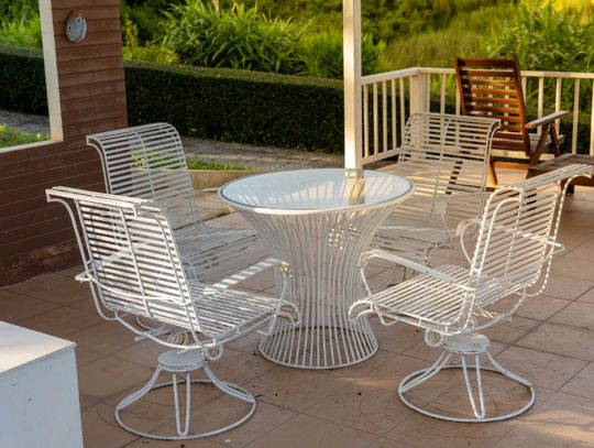 A white metal patio table and four matching chairs on a covered porch, with greenery and wooden seating in the background.