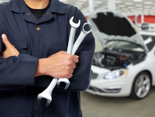 A person in a dark work uniform holding two wrenches in front of a car with an open hood inside a repair shop.