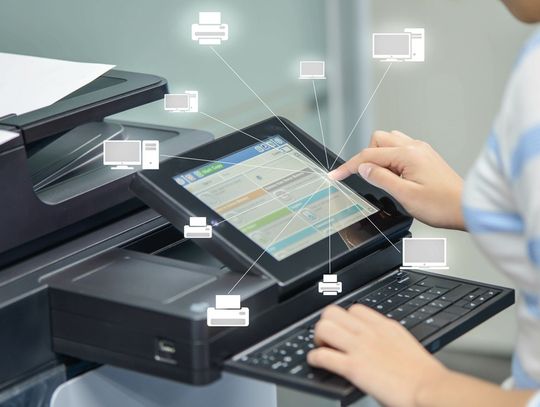 A woman presses buttons on an office printer with a stack of papers. A glass wall is visible in the background.