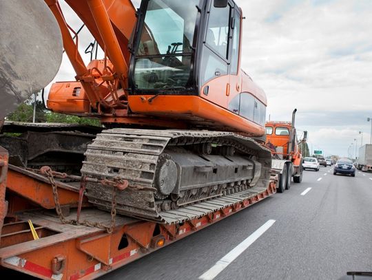 A heavy-duty semi-trailer drives down the highway, transporting an orange construction bulldozer next to other cars.