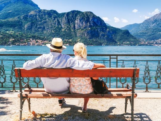 Couple sits on a lakeside bench overlooking blue water and towering green mountains under a clear summer sky.