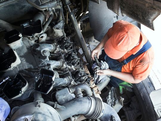Overhead view of a mechanic in an orange hat, orange shirt, and overalls working on an open truck engine.