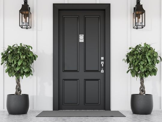 A black front door is situated between small potted trees in black pots and lights attached to the white siding.