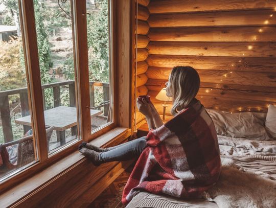 Woman wrapped in a red plaid blanket enjoys a warm drink beside large windows overlooking trees in a cozy log cabin.